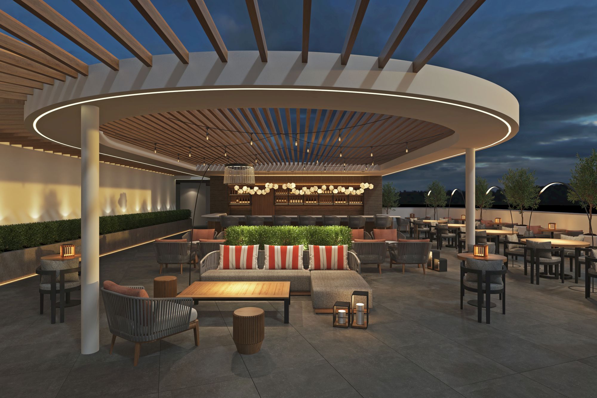 A modern outdoor rooftop lounge with a circular overhead structure, cozy seating with striped cushions, multiple tables, and a bar area in the distance at dusk.