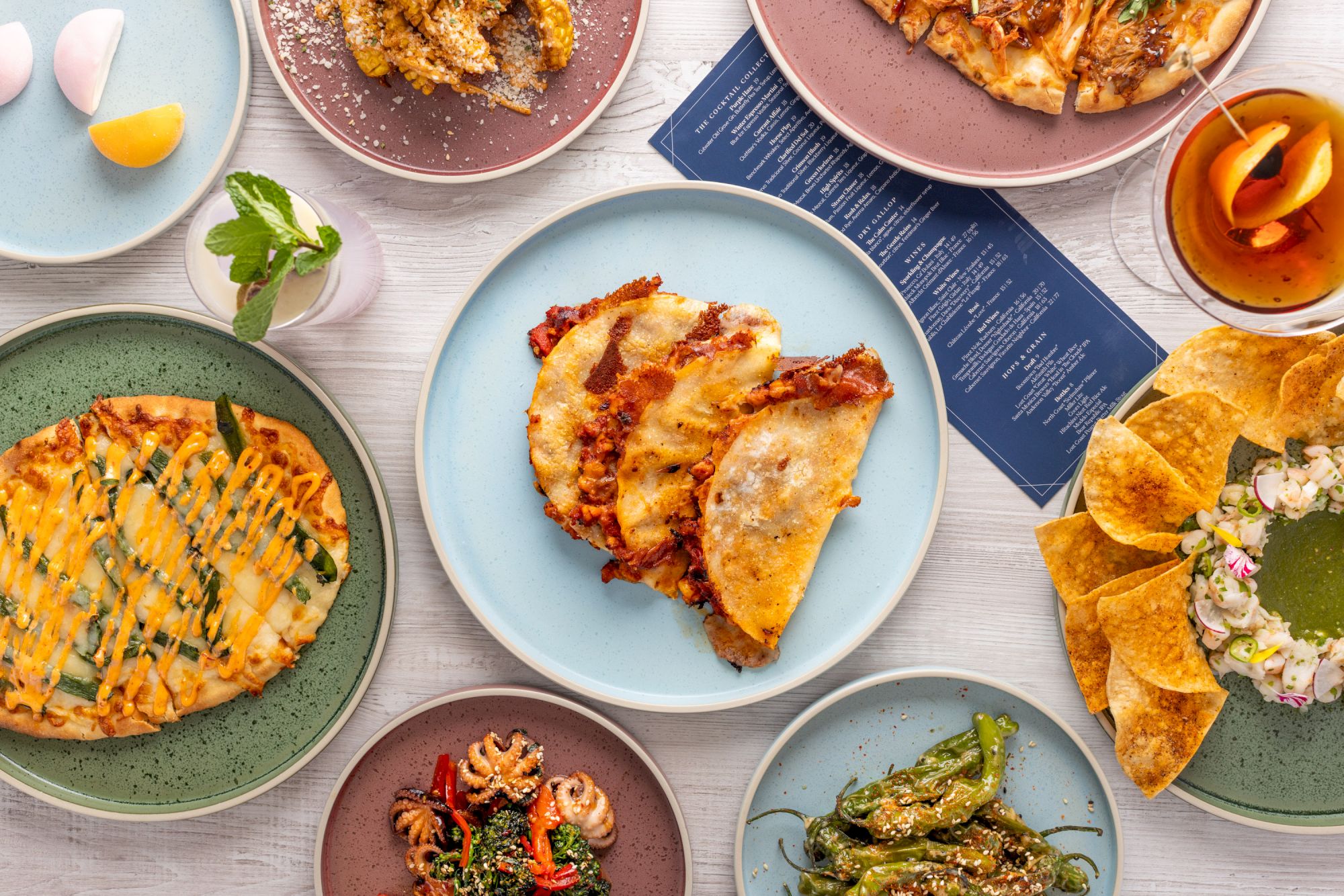 A vibrant spread of assorted dishes: pies, quesadillas, tacos, greens, and colorful plates, arranged around a rustic table.