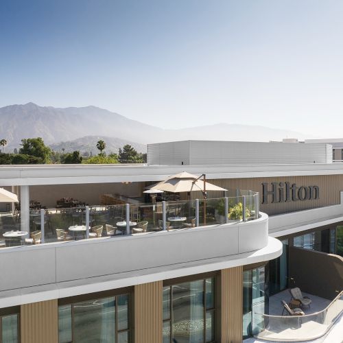 A modern hotel terrace with umbrellas and diners, the Hilton logo visible, set against palm trees and mountain views.
