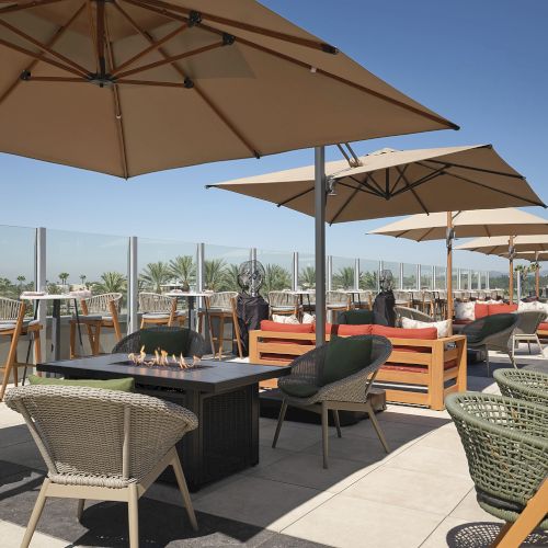 Sunny outdoor patio with umbrellas, woven chairs, a dining table, and a built-in fire pit beside palm trees.
