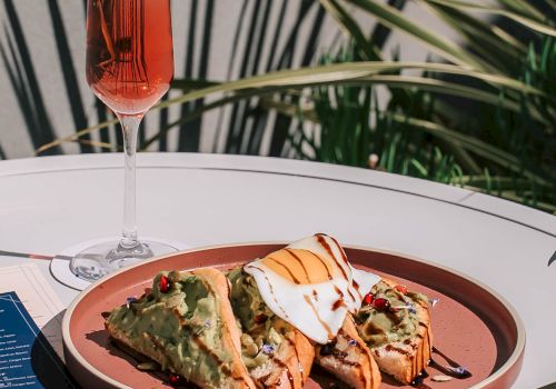 A fancy plate with savory bites, a glass of pink drink, and a blue menu on a white table. Bold flavors ahead.