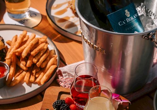 A champagne bottle chilling in a bucket, plus assorted snacks: fries, a veggie-and-fruit board with berries, and two colorful cocktails on a wooden table.