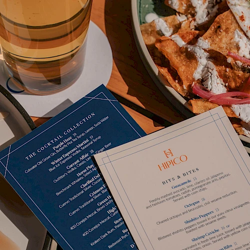 A beer glass beside a plate of nachos and two menus on a table.
