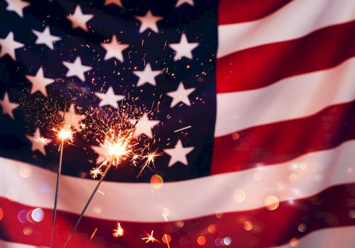 A burst of fireworks lights up a backdrop of the American flag with stars and glittering bokeh in the foreground.