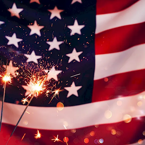 A burst of fireworks lights up a backdrop of the American flag with stars and glittering bokeh in the foreground.